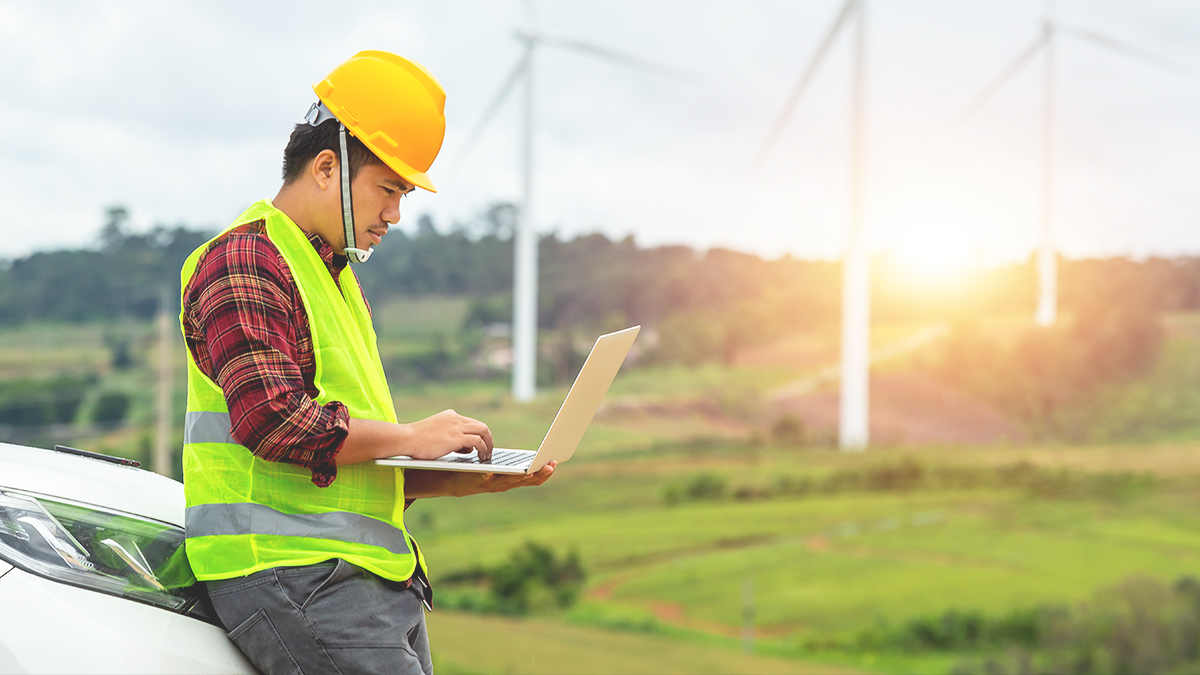 Ein Arbeiter in einer gelben Sicherheitsweste und einem gelben Schutzhelm benutzt einen Laptop, während er sich gegen die Motorhaube eines Autos in der Nähe eines Windparks lehnt