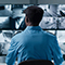 A security officer wearing a blue uniform shirt and cap watching a large spread of wall-mounted security camera displays